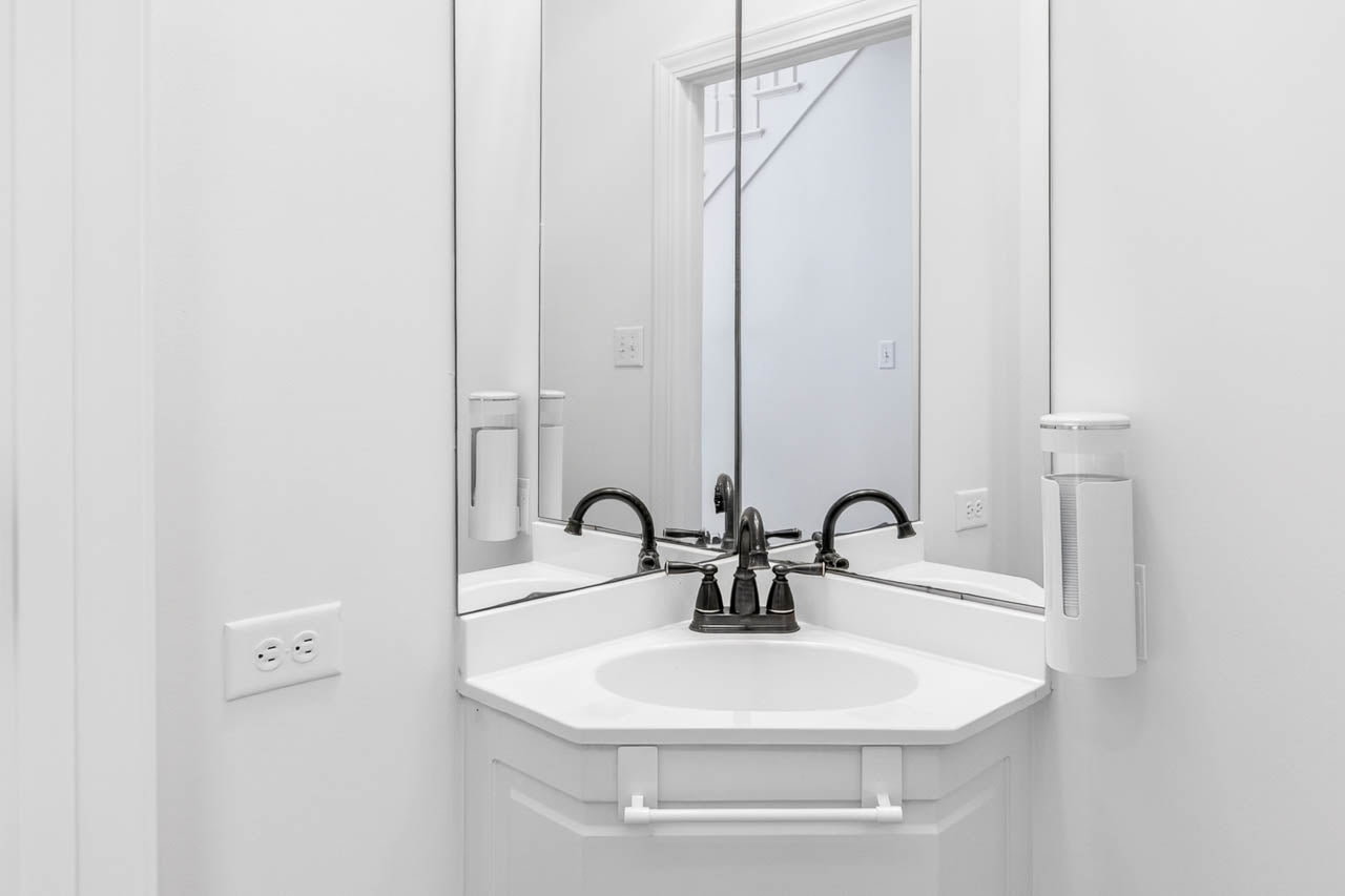A white corner bathroom sink with a black faucet is positioned beneath two mirrors that meet at a right angle. There is a towel rack beneath the sink and a wall-mounted soap dispenser on the right side.