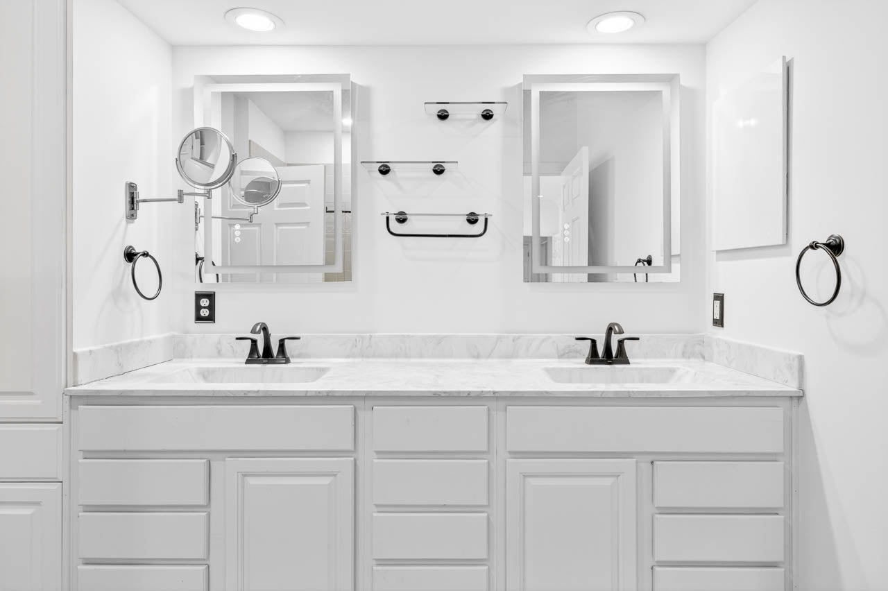 A modern white bathroom with a double sink vanity, two mirrors, black faucets, and black towel rings. The countertop is marble, and the lighting is recessed. Shelves and a towel bar are mounted between the mirrors.