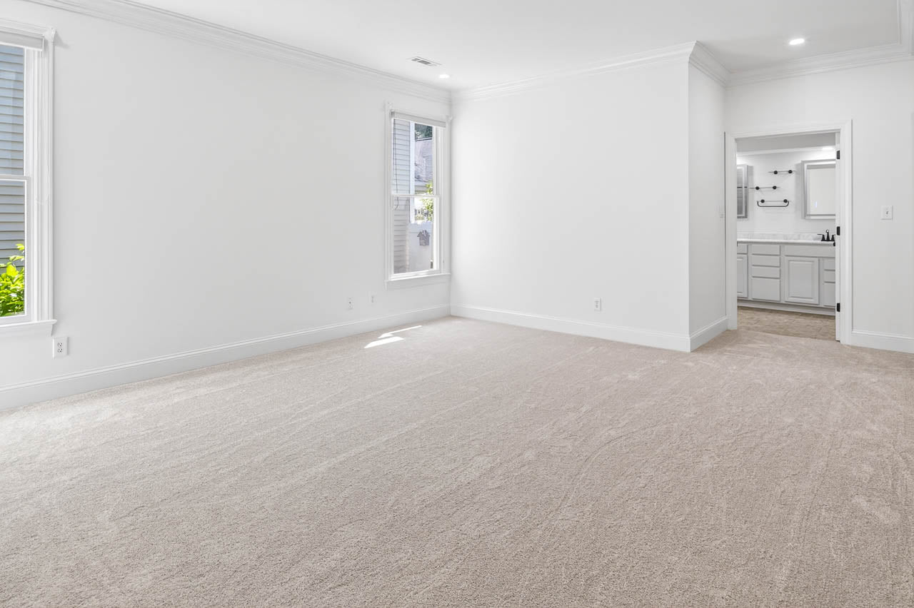 A spacious, empty room with light beige carpet, white walls, two tall windows on the left, and an open doorway leading to a bathroom with double sinks and a large mirror.
