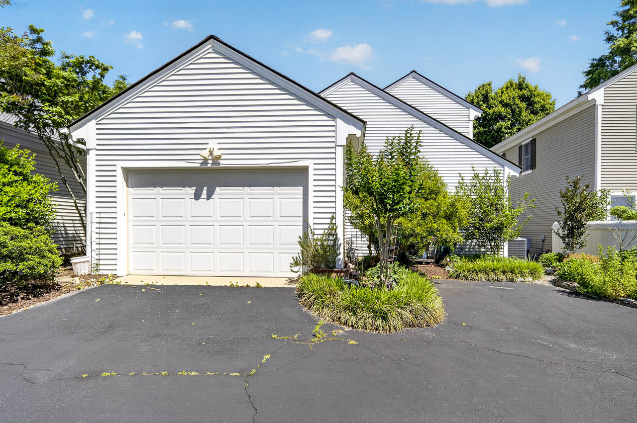 Single-story house with white siding, a two-car garage, blacktop driveway, and small landscaped garden in front, surrounded by bushes and trees under a blue sky with scattered clouds.
