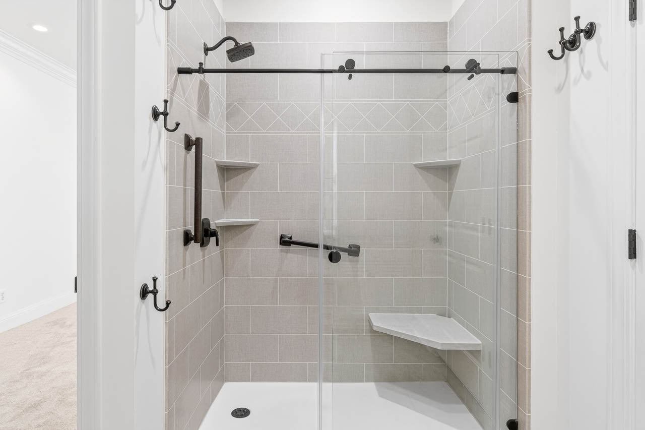 Modern walk-in shower with light gray tiles, black fixtures, glass doors, two built-in corner shelves, a grab bar, and a white corner bench, viewed from the bathroom entrance.