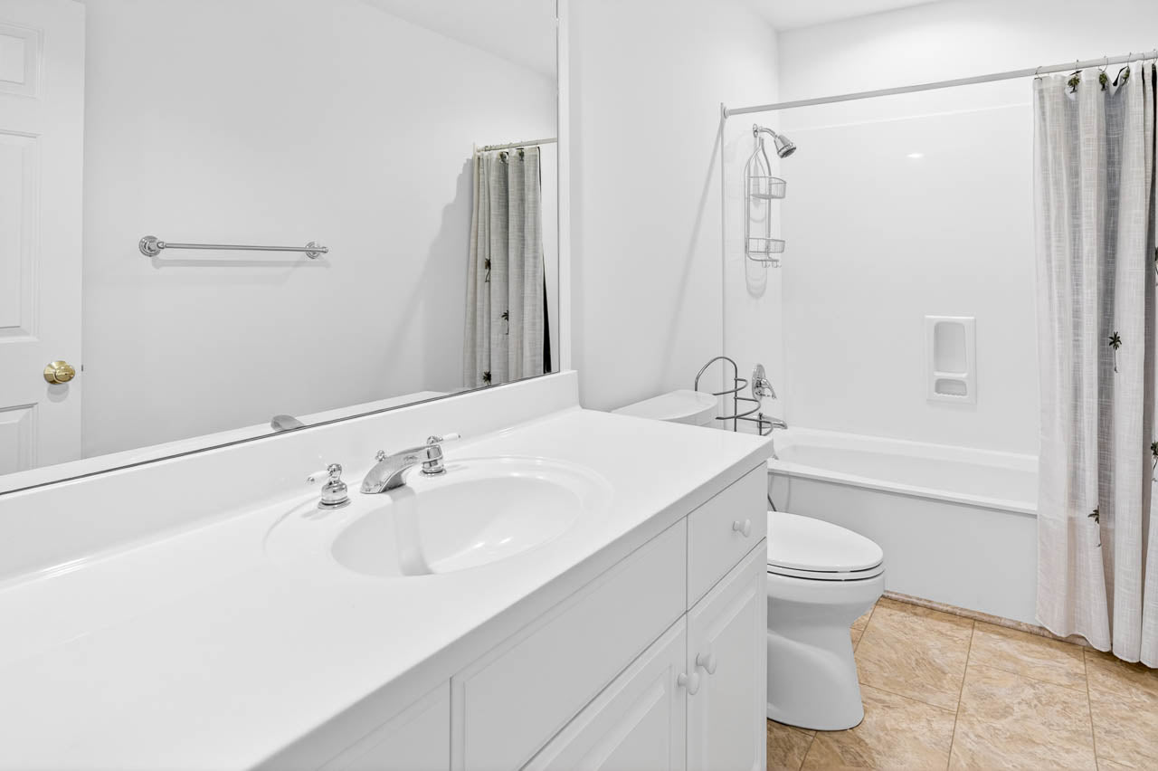 A clean, white bathroom with a large mirror, single sink vanity, toilet, and a bathtub with a shower curtain. The floor is tiled, and the room has simple, minimalistic decor.