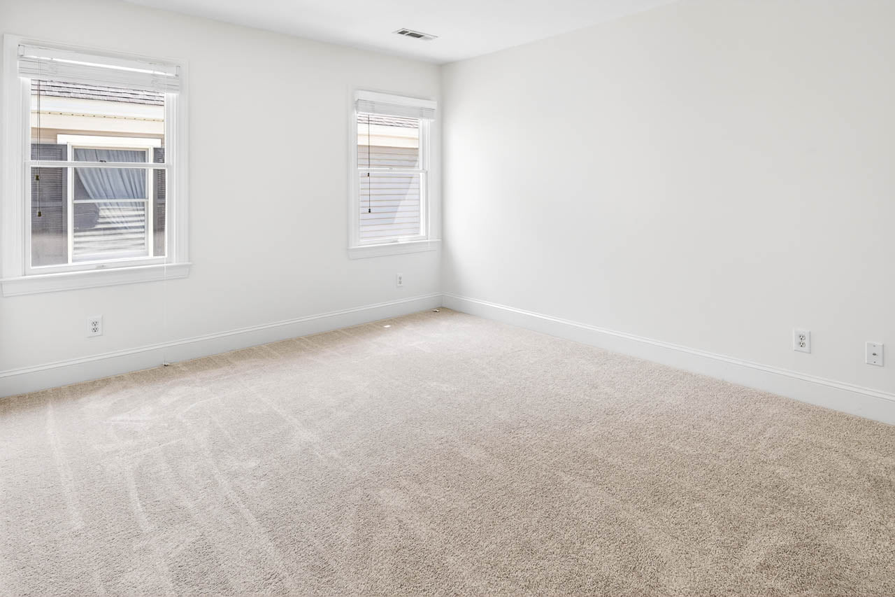 A bright, empty room with light beige carpet, white walls, and two windows letting in natural light. The space appears clean and spacious, suitable for use as a bedroom or office.