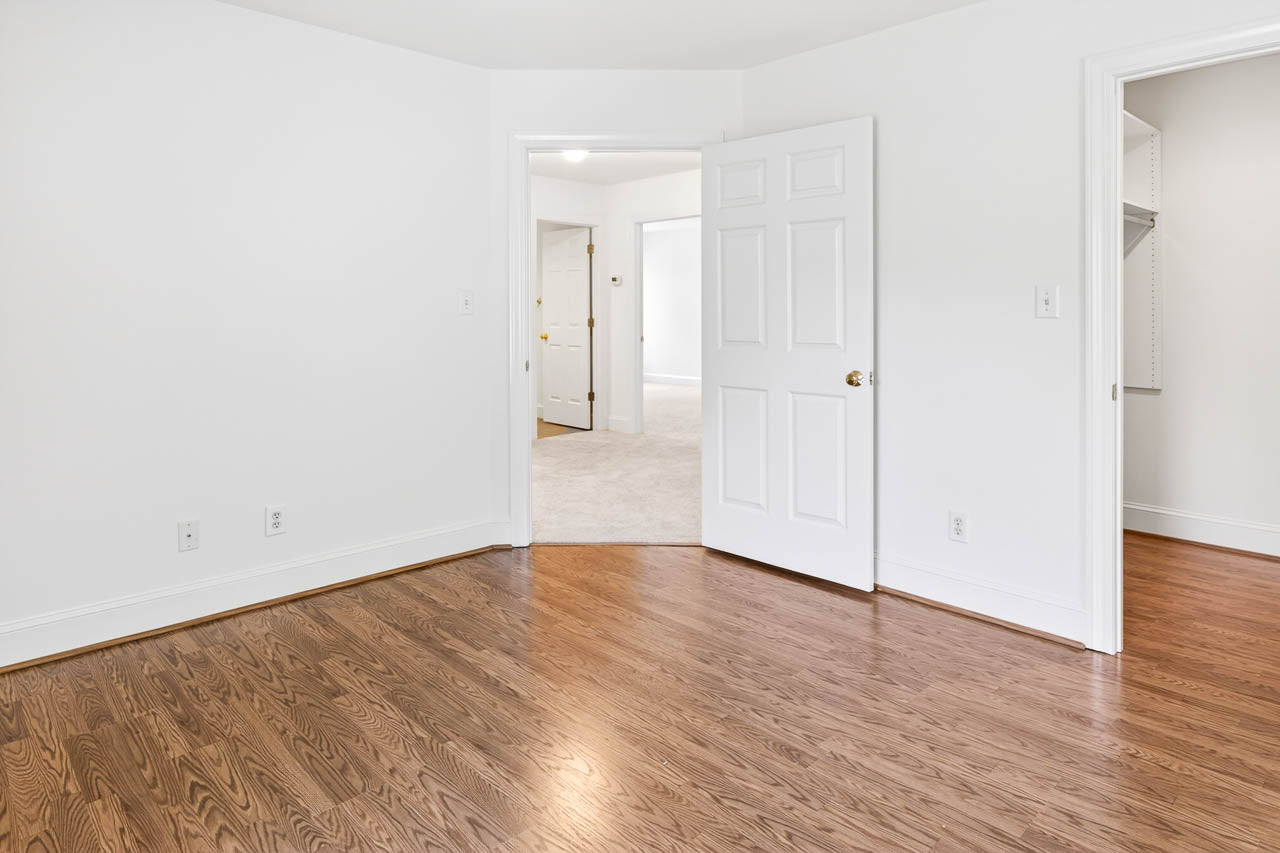 Empty room with hardwood floors, white walls, and two open doors leading to carpeted areas and a walk-in closet with built-in shelves. The space is well-lit and minimal, with visible wall outlets.