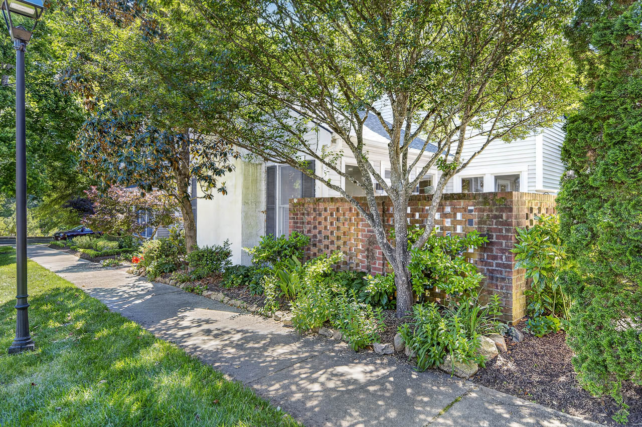 A small brick patio with lattice-style walls is surrounded by lush green plants and shaded by leafy trees next to a sidewalk and a white house in a suburban neighborhood.