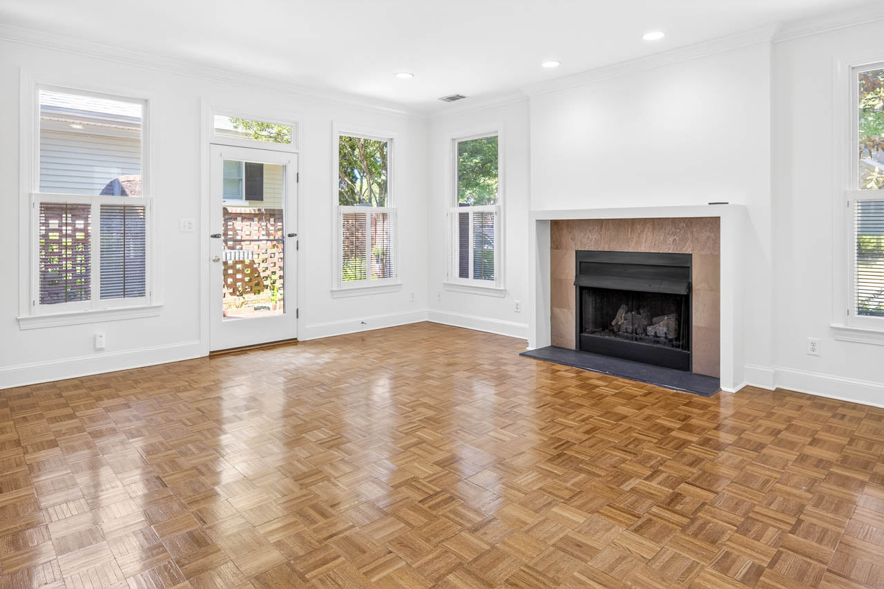 Bright, empty living room with large windows, a glass door leading outside, a wood parquet floor, and a fireplace with a simple mantel. White walls and ceiling create a clean, open atmosphere.