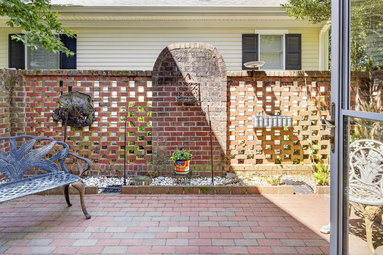 A small brick patio with a decorative brick wall, metal bench, potted plants, garden art, and a sunlit area. The wall features open brickwork and mounted decorations, with white gravel below and a house in the background.