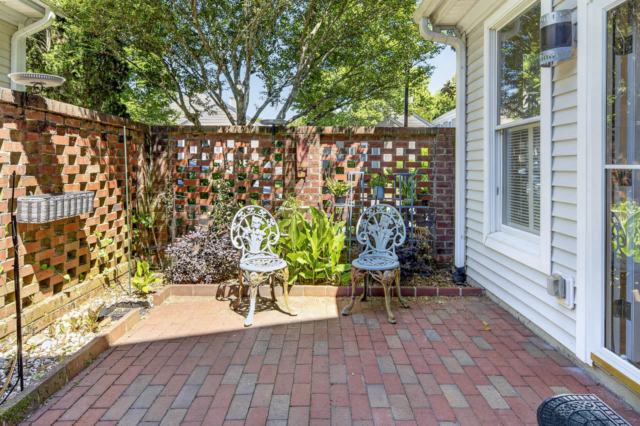 A small brick patio with two ornate metal chairs, surrounded by a brick privacy wall with lattice design, lush green plants, and a white house exterior with windows. Sunlight filters through trees overhead.