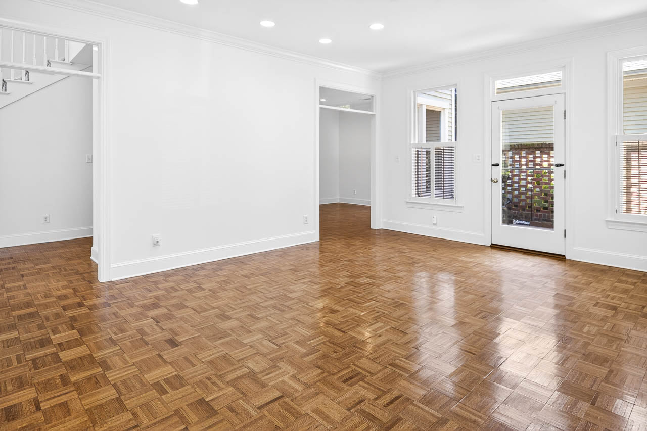 Bright, empty living room with polished parquet wood flooring, white walls, recessed ceiling lights, large windows, a glass door, and a staircase partially visible on the left.
