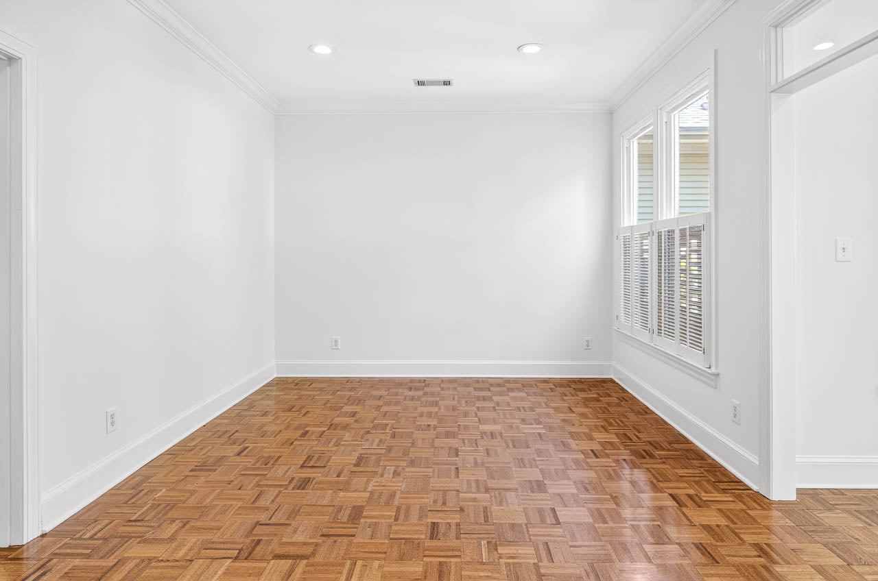 Bright, empty room with white walls, parquet wood flooring, recessed ceiling lights, and large windows with white blinds letting in natural light. Doorways are visible on the right side of the room.