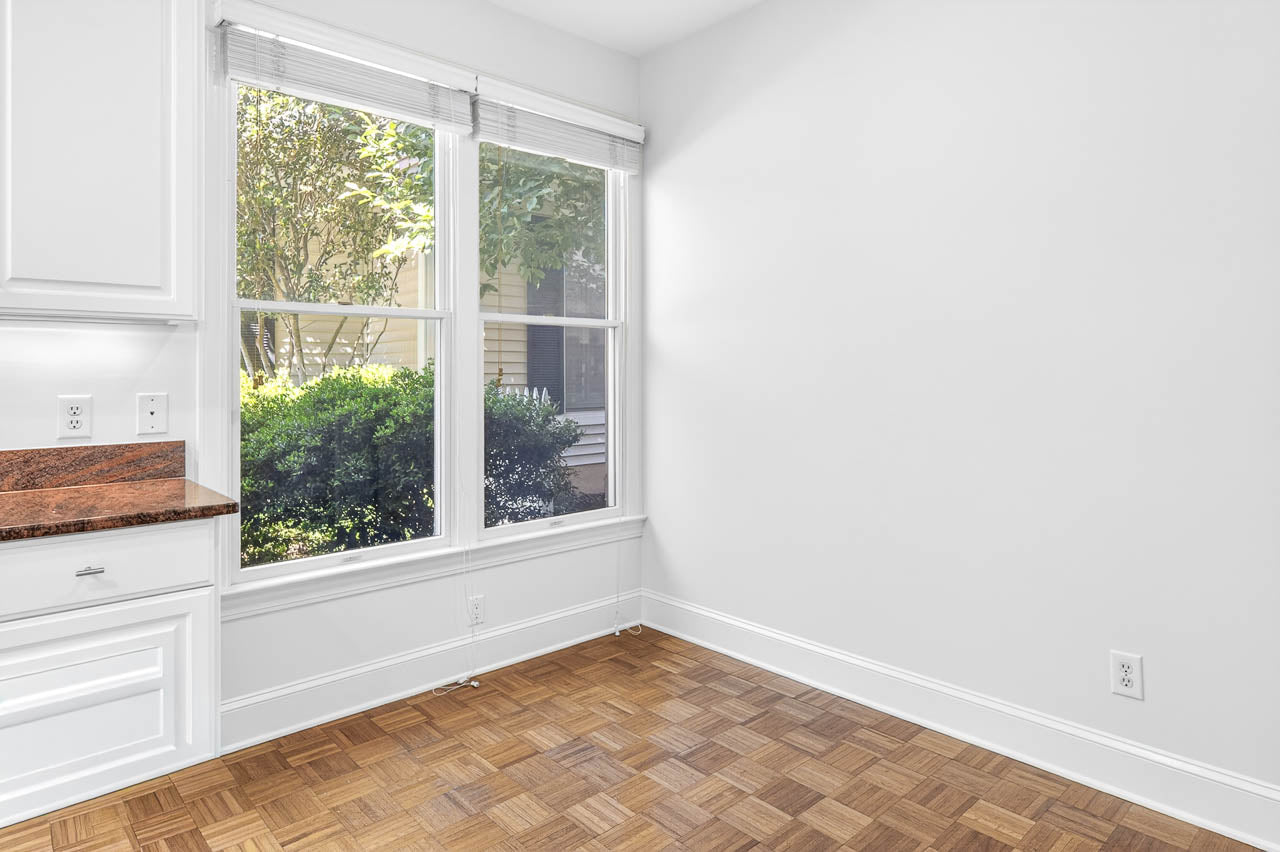 A bright, empty room with white walls, wood parquet flooring, and large windows letting in natural light. Green shrubs and part of a house exterior are visible through the windows. Part of a white kitchen counter is on the left.