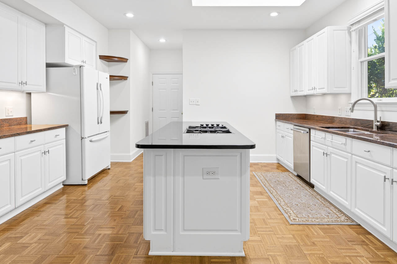 Bright, modern kitchen with white cabinets, wood flooring, a large island with a cooktop, stainless steel appliances, brown countertops, open shelves, and a window above the sink letting in natural light.