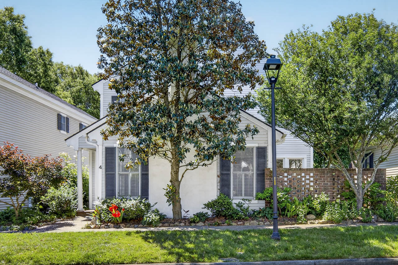 A small white house with gray shutters is partly shaded by a large tree in the front yard. A brick wall, garden beds, and a black lamppost are visible, with bright green grass and other trees surrounding the home.