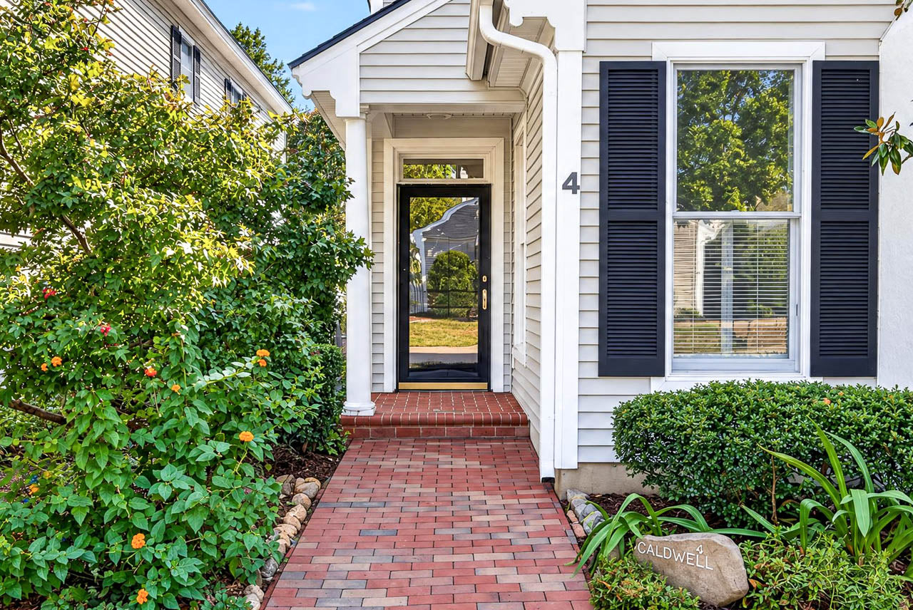 Front entrance of a house with a brick walkway leading to a glass-paneled door, black window shutters, and a decorative stone engraved with the name “Caldwell.”