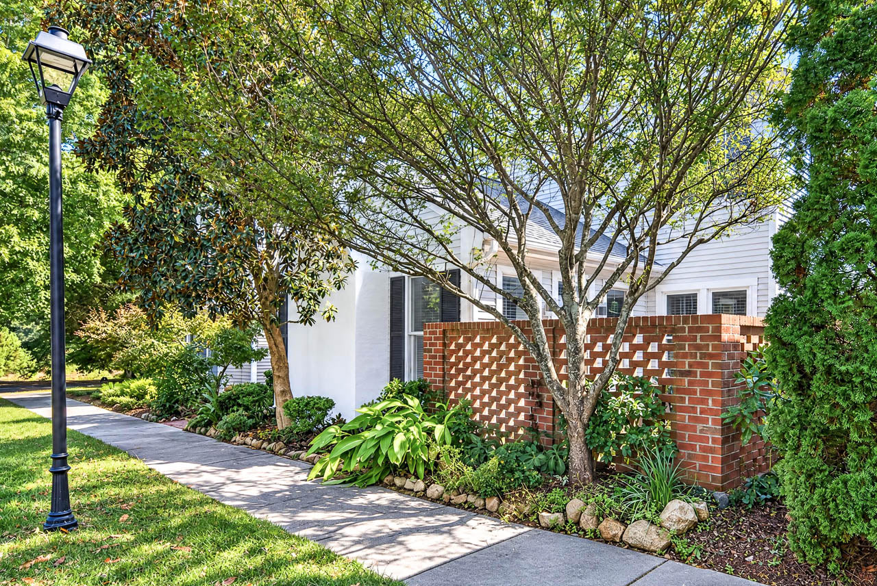 A sidewalk lined with a street lamp leads past a white house with a decorative brick privacy wall and large trees.
