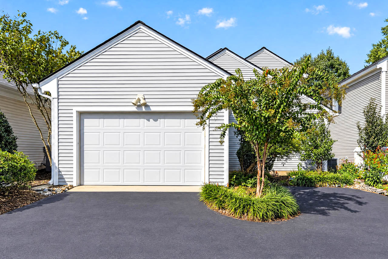 A suburban house with a white garage door, light grey siding, and a paved driveway with a small garden island.