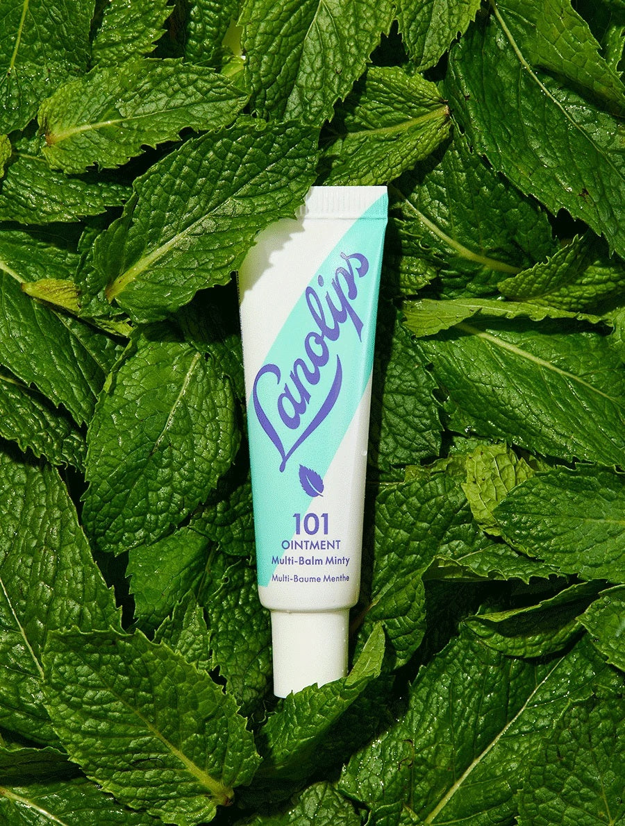 A woman holds a tube of LANOLIPS - 101 OINTMENT MULTI-BALM MINTY near her lips, dressed in a cream sweater against a blue background—highlighting LANOLIPS’ perfect minty hydration for dry lips.