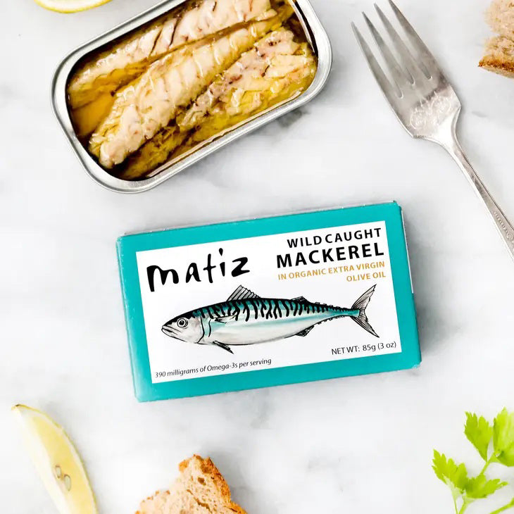 The opened tin of MATIZ - Wild Caught Mackerel in Organic Olive Oil reveals tender fish slices, with an unopened package featuring a mackerel illustration beside it. A fork, crusty bread, and lemon slices are elegantly arranged on a pristine white background. Made in Spain by MATIZ ESPANA with care.
