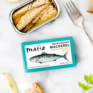 The opened tin of MATIZ - Wild Caught Mackerel in Organic Olive Oil reveals tender fish slices, with an unopened package featuring a mackerel illustration beside it. A fork, crusty bread, and lemon slices are elegantly arranged on a pristine white background. Made in Spain by MATIZ ESPANA with care.