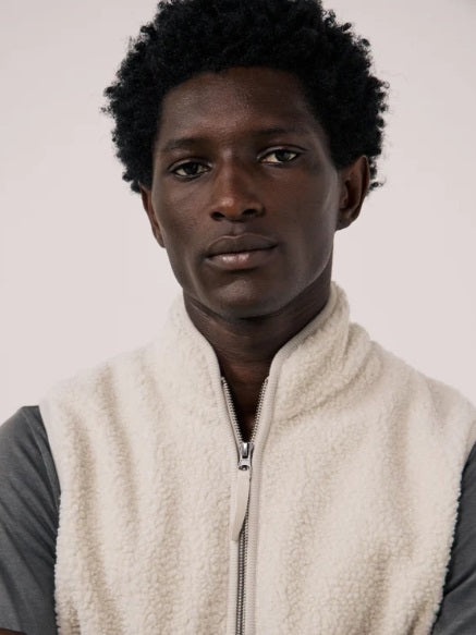 A man is wearing the HARTFORD - BASTA SHERPA VEST IN NATURAL over a gray long-sleeve shirt, holding the vest’s zipper with one hand. The plain, light-colored background complements the cozy, Hartford-inspired style of this men’s vest from HARTFORD.