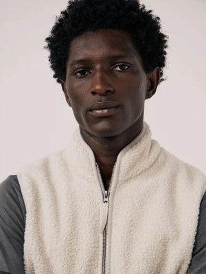 A man is wearing the HARTFORD - BASTA SHERPA VEST IN NATURAL over a gray long-sleeve shirt, holding the vest’s zipper with one hand. The plain, light-colored background complements the cozy, Hartford-inspired style of this men’s vest from HARTFORD.