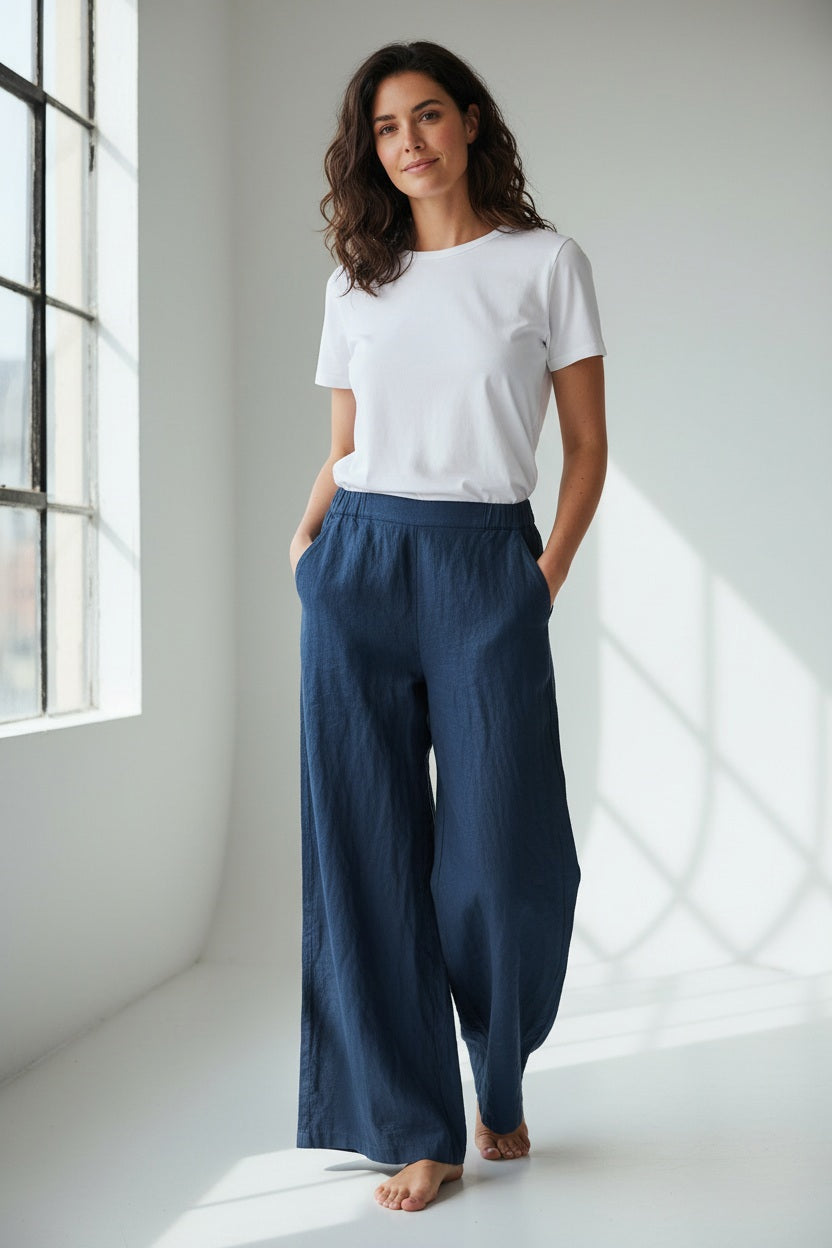 A woman stands barefoot in a bright room, wearing a white cotton T-shirt and the CP SHADES Wendy Palazzo Pant, with its loose high-rise wide-leg silhouette, hands in her pockets and a relaxed expression.