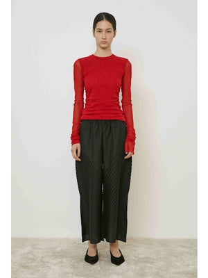 A woman with long dark hair in a low ponytail stands in profile against a plain light background, wearing the MEIMEIJ MESH LAYERING TOP by MEIMEIJ in red and a black skirt for a minimalist Italian fashion look.