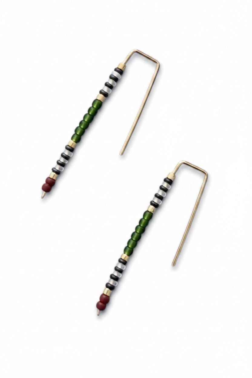 The ALICE RISE - BIG QUILL EARRINGS IN FOREST GREEN by ALICE RISE feature 14k gold-filled hooks and small red, green, black, and white beads. Displayed parallel on a textured light blue background, their hooks face opposite directions.