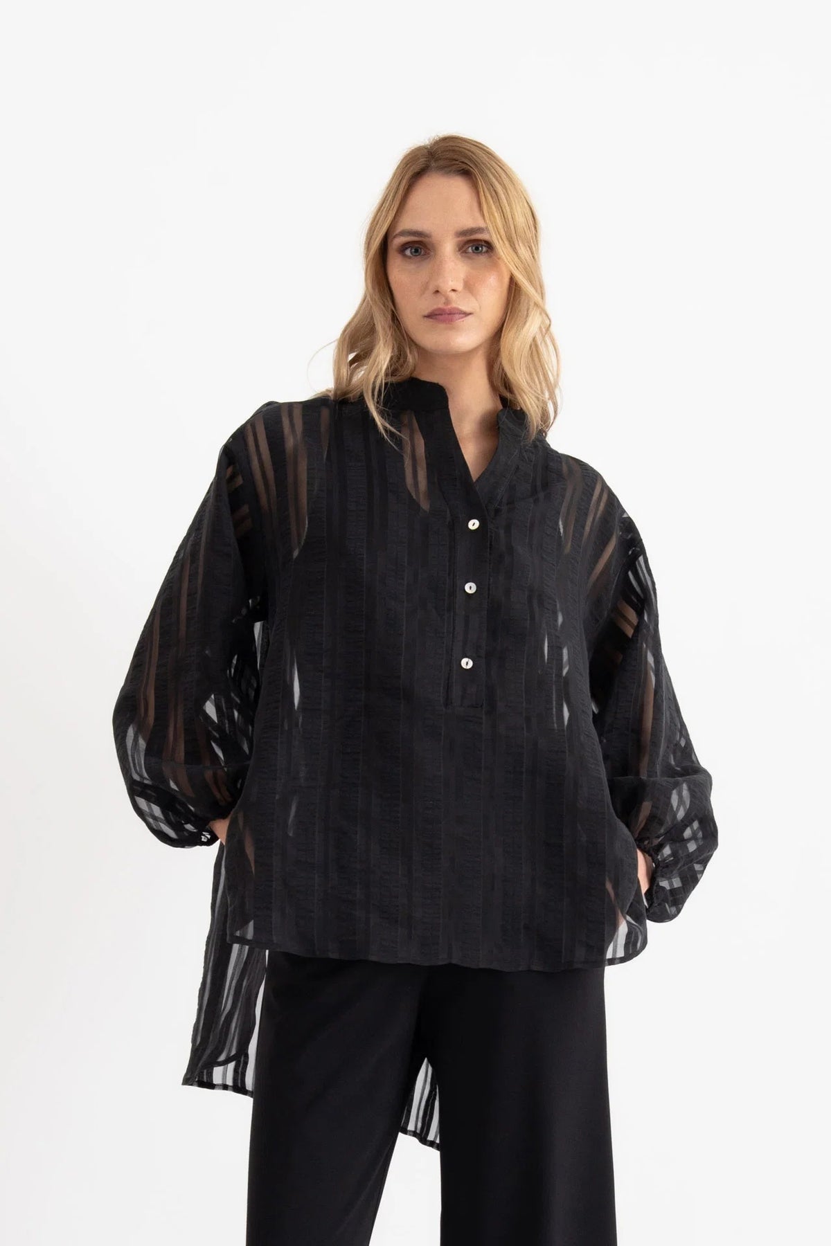 A woman stands in a bright room, looking at the camera with a neutral expression. She is wearing the VICARIO CINQUE SEAN SHEER STRIPE SHIRT, a black sheer long-sleeve blouse crafted from lightweight fabric with a band collar by VICARIO CINQUE, paired with black wide-leg pants and black sandals.