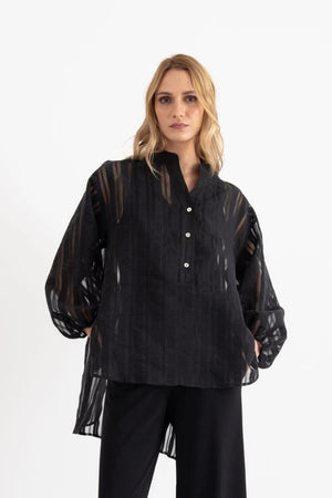 A woman stands in a bright room, looking at the camera with a neutral expression. She is wearing the VICARIO CINQUE SEAN SHEER STRIPE SHIRT, a black sheer long-sleeve blouse crafted from lightweight fabric with a band collar by VICARIO CINQUE, paired with black wide-leg pants and black sandals.