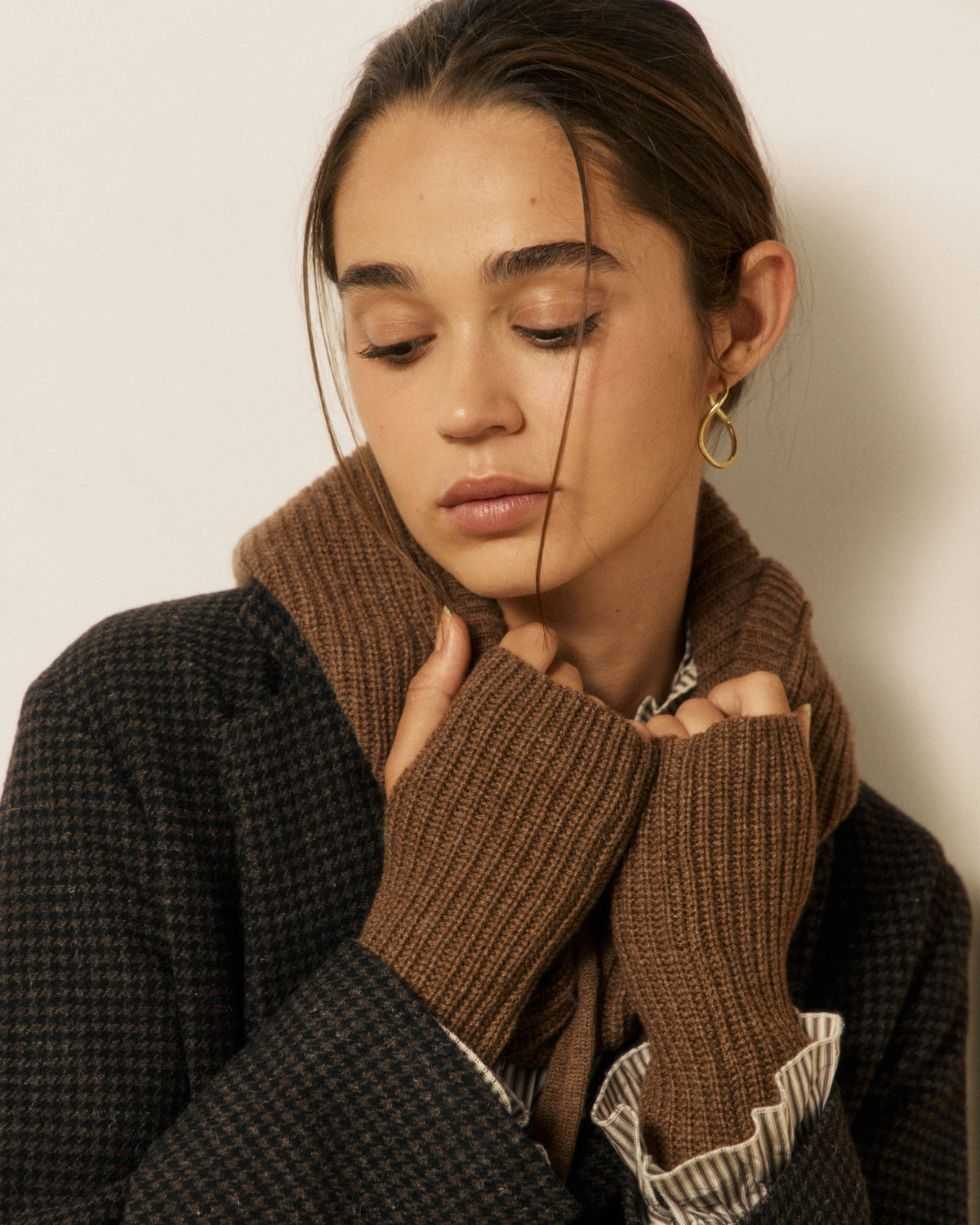 A woman in a dark textured coat and brown knit scarf gazes thoughtfully downward, her hands near her face showing off the elegant HARTFORD - MESSIA Fingerless Mittens in Cognac. A gold teardrop earring completes her ensemble of timeless autumn-winter accessories.