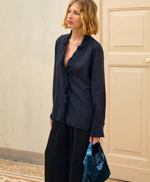 A person with shoulder-length blonde hair is wearing the MOMONI - Marc Blouse in textured cotton silk, which features ruffles along the collar and cuffs. They are also wearing black wide-leg pants and holding a teal-colored clutch. The background includes a light-colored door and wall.