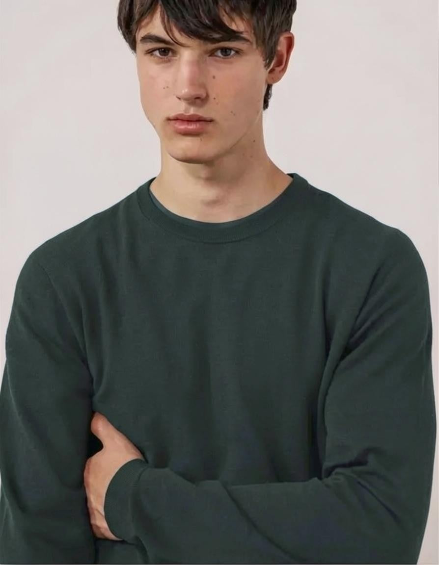 A young man stands with his hands in his pockets against a white background, wearing the HARTFORD Extra-Fine Merino Wool Crew Pullover in Forest and light-colored pants.