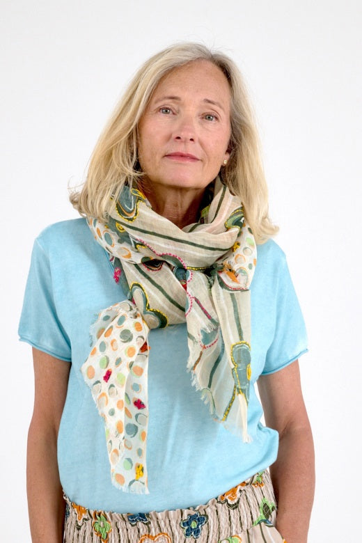 A woman with blonde hair stands against a white background, wearing the STORIATIPIC ANNE Scarf in beige featuring pastel stripes and floral prints with hand-embroidered linen details, paired with matching pants and green sandals.