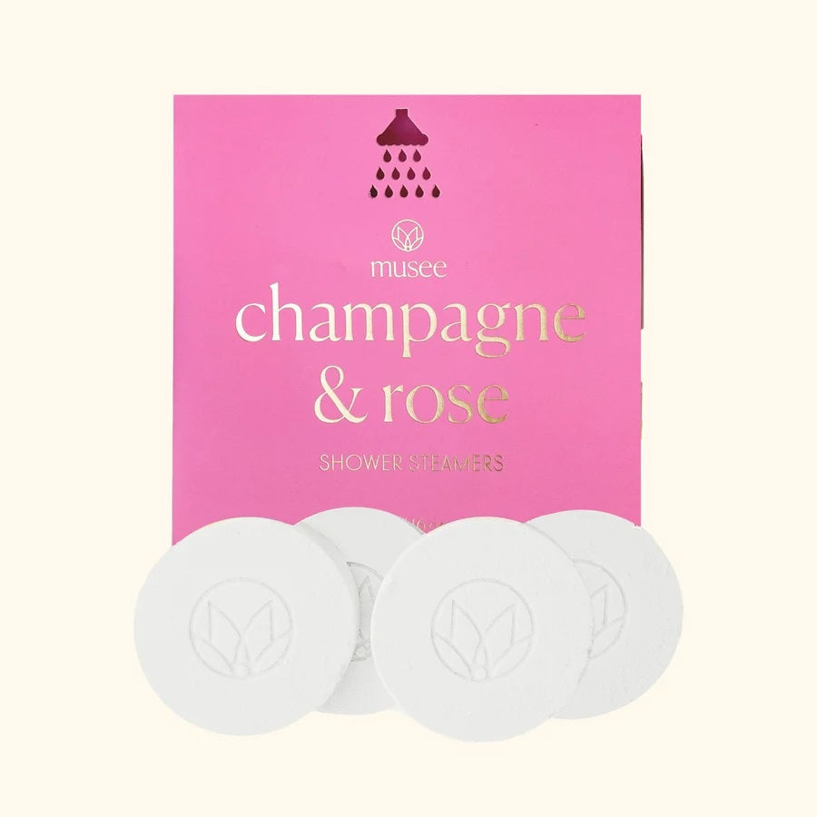 Four round white shower steamers are placed in front of a pink MUSEE package labeled “MUSEE - CHAMPAGNE & ROSE SHOWER STEAMERS” in gold and white text with a small showerhead graphic above, offering a natural bath product designed to turn your daily routine into a luxurious spa shower experience.