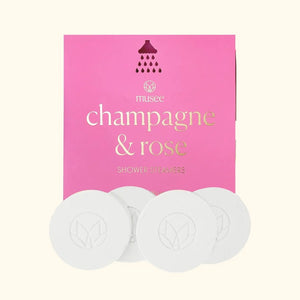Four round white shower steamers are placed in front of a pink MUSEE package labeled “MUSEE - CHAMPAGNE & ROSE SHOWER STEAMERS” in gold and white text with a small showerhead graphic above, offering a natural bath product designed to turn your daily routine into a luxurious spa shower experience.