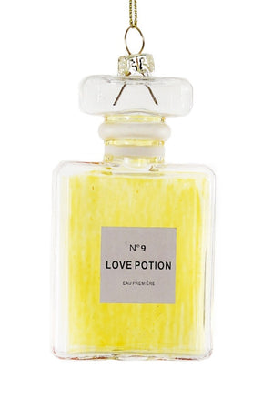 A tall glass bottle labeled "LOVE POTION ORNAMENT" from CODY FOSTER features a decorative stopper and a gold string that enhances its elegant height. Filled with a captivating yellow liquid, it promises enchantment with every glance.