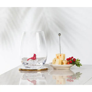 An ABBOTT CARDINAL STEMLESS WINE GLASS featuring a small red bird figure inside, sitting next to a plate of cubed cheese and holly berries.