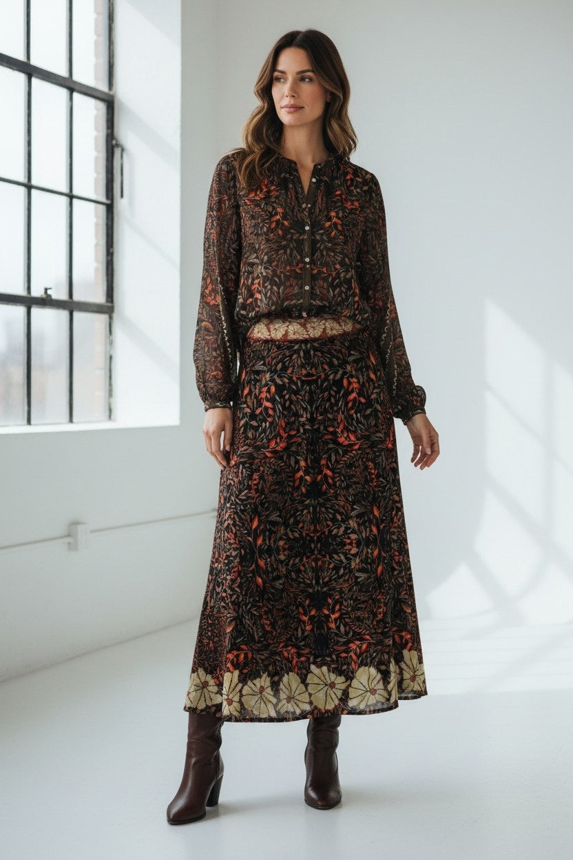 A woman stands in a bright room with large windows and white walls, dressed in a long-sleeved, patterned dress in autumn hues paired with brown heeled boots. Her look is elevated by the addition of the FRANCO FERRARI JEMMA WOOL SKIRT, featuring an elegant botanical print that infuses her ensemble with nature-inspired charm.