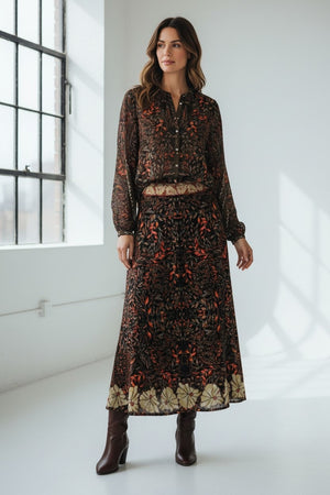 A woman stands in a bright room with large windows and white walls, dressed in a long-sleeved, patterned dress in autumn hues paired with brown heeled boots. Her look is elevated by the addition of the FRANCO FERRARI JEMMA WOOL SKIRT, featuring an elegant botanical print that infuses her ensemble with nature-inspired charm.