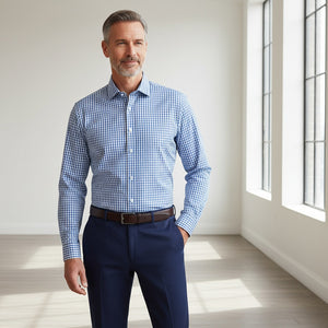 A man wearing the COLLARS AND CO Semi Spread Collar Apex Performance Dress Shirt in Vincenzo Blue Tattersall walks confidently down a modern office hallway, with large windows, plants, and people working in the background.