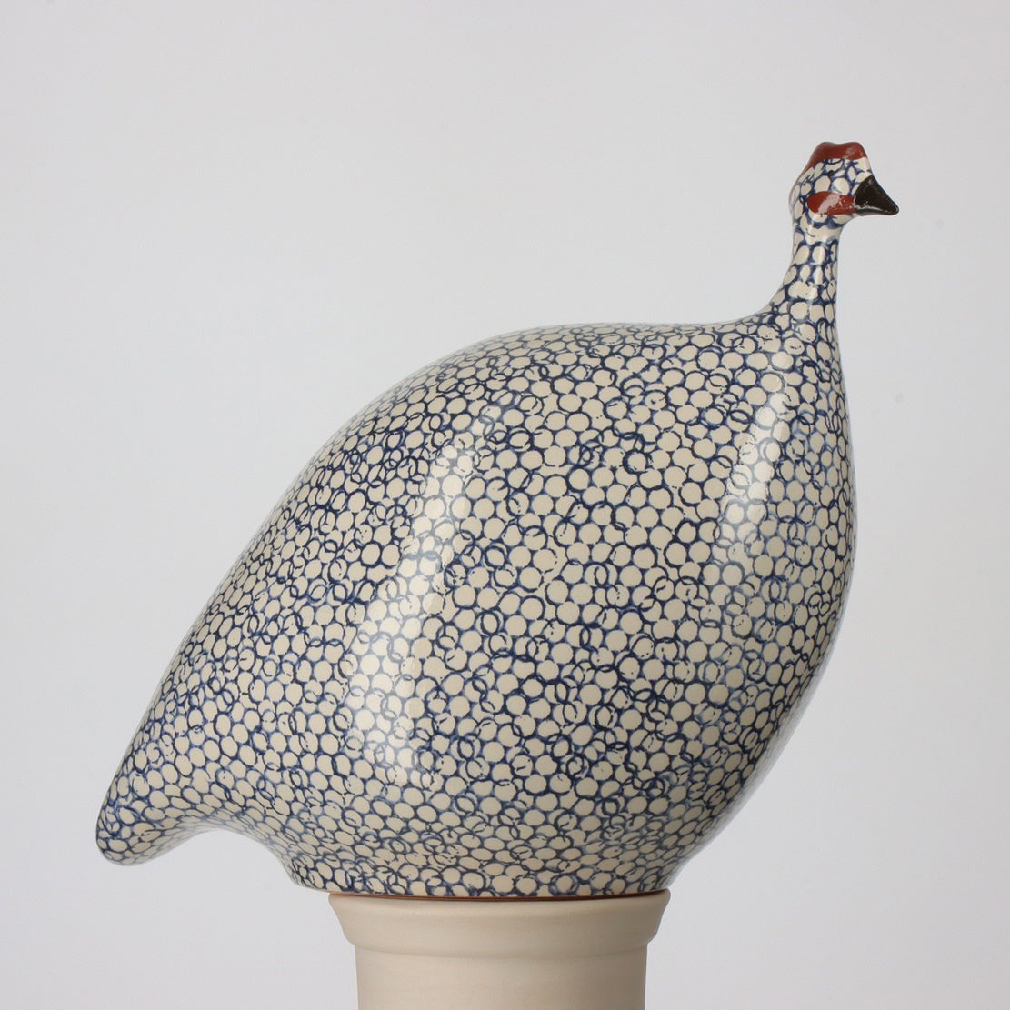 The LES CERAMIQUES DE LUSSAN - MEDIUM CERAMIC FRENCH GUINEA IN WHITE SPOTTED COBALT is a handcrafted pottery sculpture of a French guinea hen, featuring a speckled blue and white design, and it elegantly rests on a cream-colored pedestal against a simple backdrop.