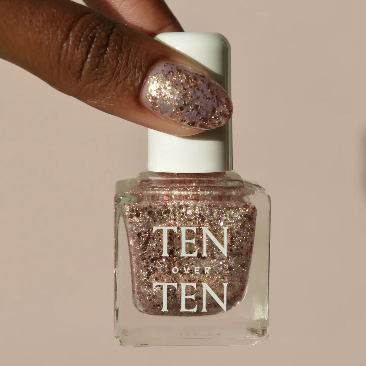 A glass bottle of TENOVERTEN - NAIL POLISH from the brand TEN OVER TEN, featuring a white cap and a dusty pink nail color. The front label displays "TEN OVER TEN" in white uppercase letters. This vegan, eight-free formula ensures both beauty and peace of mind. The background is plain and light-colored.
