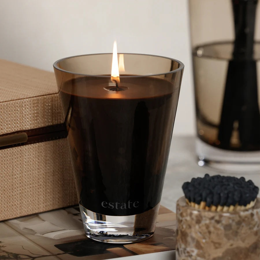 The ZODAX Thistle Estate Candle (10.5 oz) in black is displayed in a glass holder beside its matching box, both reflecting the fresh California coast fragrance.