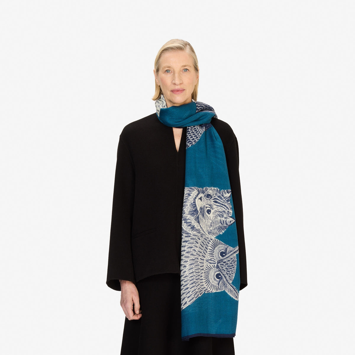 A person models the INOUI EDITIONS - OWLS ON A BRANCH WOOL SCARF IN BLUE by INOUI, featuring intricate white animal and plant motifs, with only their upper face and torso visible against a plain white background.