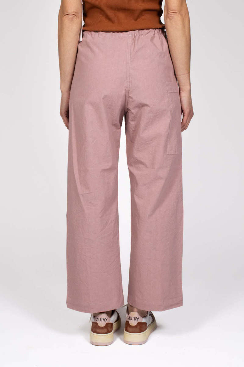 A person stands against a plain background wearing THE LITTLE PROJECT&#39;s AMBER PANT in pink, crafted from Japanese cotton poplin with a drawstring waist and subtle contrast stitching, paired with a sleeveless brown top and white and brown sneakers.