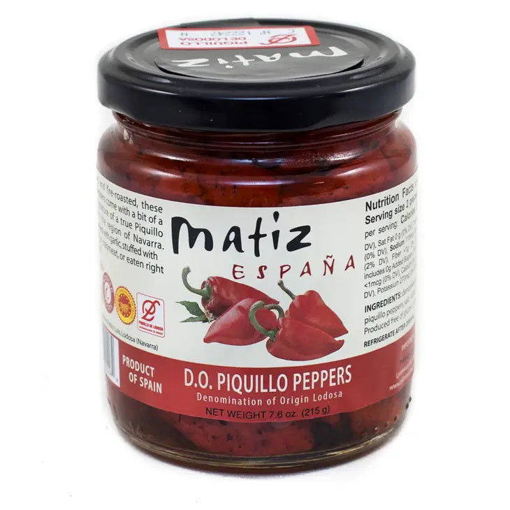 A jar of MATIZ - PIQUILLO PEPPERS from MATIZ ESPANA features a label adorned with red peppers and the phrase "Product of Spain." Weighing 7.6 oz (215 g), the label also provides nutrition facts, detailed descriptions, and an illustration of two red peppers. These hand-processed and roasted peppers deliver an intensely sweet and pungent flavor.