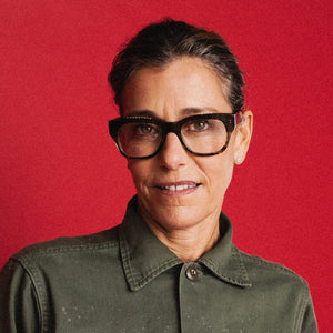 A person wearing CADDIS - MIKLOS READERS IN MADRAS PATCHWORK glasses with thick-rimmed, multicolored patterned frames against a solid red background.
