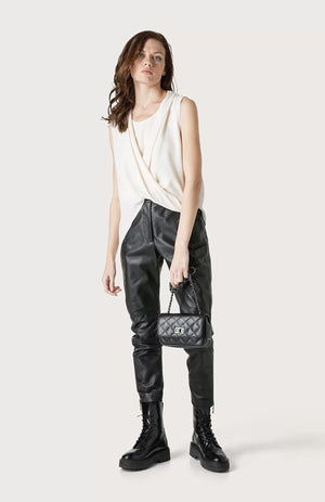 A woman with wavy brown hair stands against a plain background. She is wearing the SEVENTY Jersey Sleeveless with Draped Hemline, black leather pants, and black combat boots. She holds a small black quilted handbag by her side. Her expression is neutral.