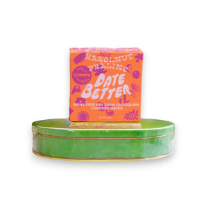 The "DATE BETTER - HAZELNUT PRALINE COVERED DATES" from DATE BETTER SNACKS features orange packaging with bold pink and purple text, adorned with abstract flower and leaf patterns. This 85% dark chocolate Medjool date treat weighs 1.8 oz.
